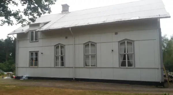A white building with a corrugated metal roof and several windows, set against a backdrop of trees and a cloudy sky.