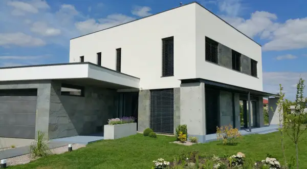 Modernt hus med vit, grå putsad fasad och stenbeklädda partier. Grönskande trädgård och garage.