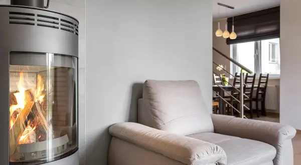 En modern kamin med brinnande ved. En bekväm fåtölj står bredvid, perfekt för en mysig kväll. Kanske dags att renovera kakelugn för en liknande känsla?