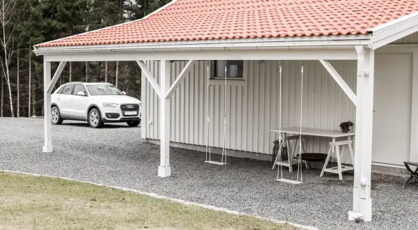 Vit bil parkerad vid en vit träfasad med röd tegeltak. En utbyggnad hus långsida med hängande gungor och ett bord.
