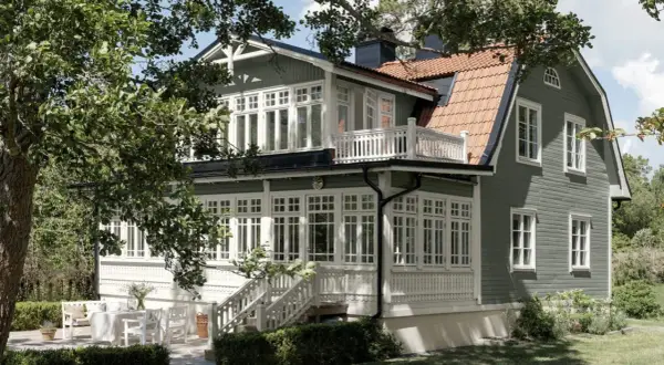 Ett charmigt grått hus med svarta knutar, omgivet av grönska. Ett vackert hus med många fönster och en veranda.