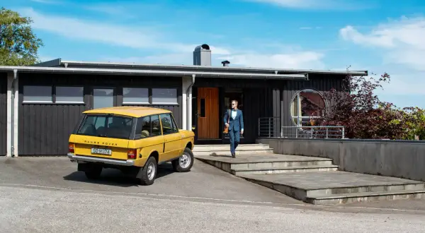 Man i kostym går mot ett funkishus från 60-talet med en gul Range Rover parkerad utanför.
