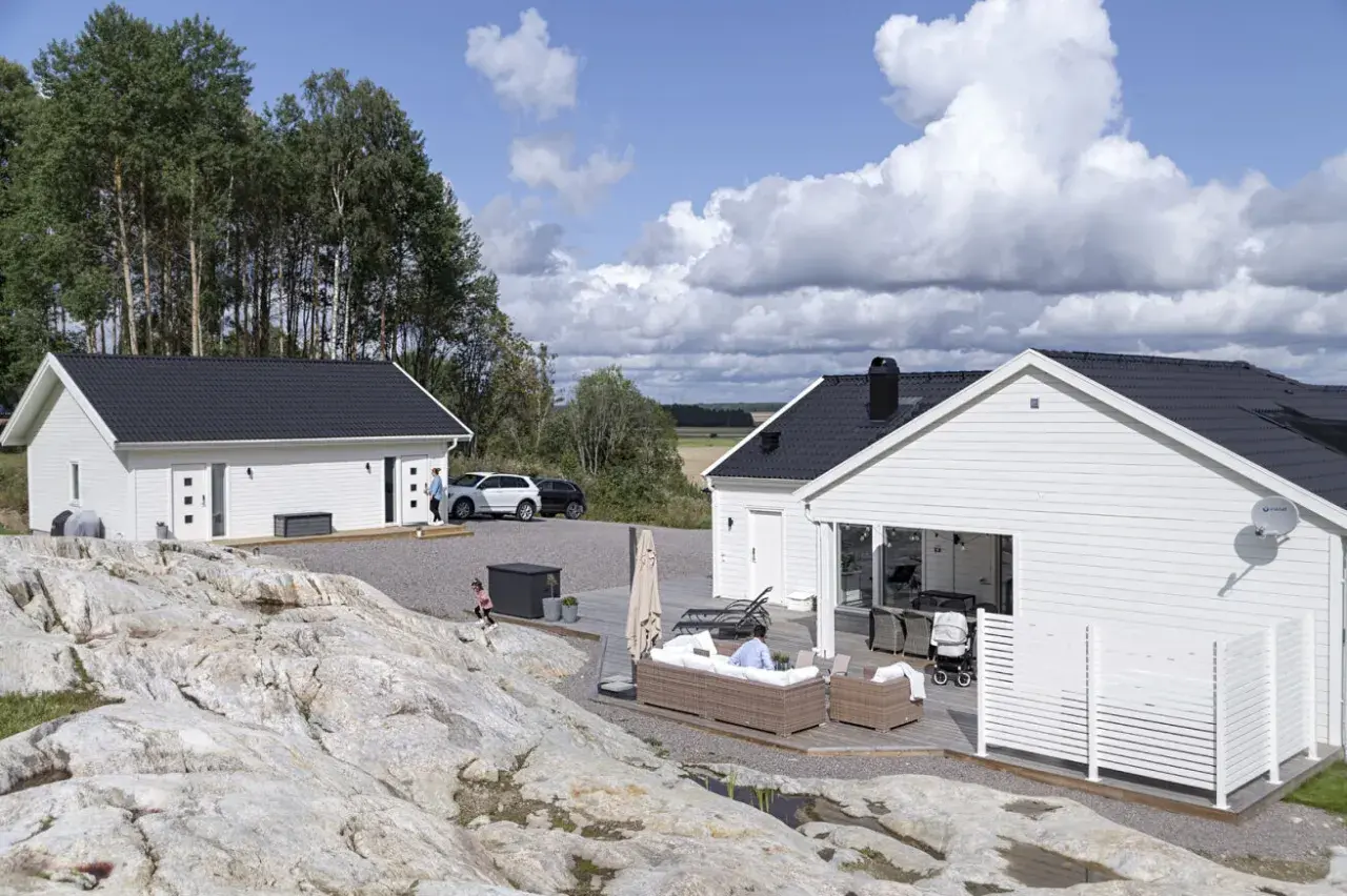 Två vita hus med svarta tak, en del av ett projekt för att bygga hus. En person sitter på en soffa utomhus, en barnvagn står bredvid.