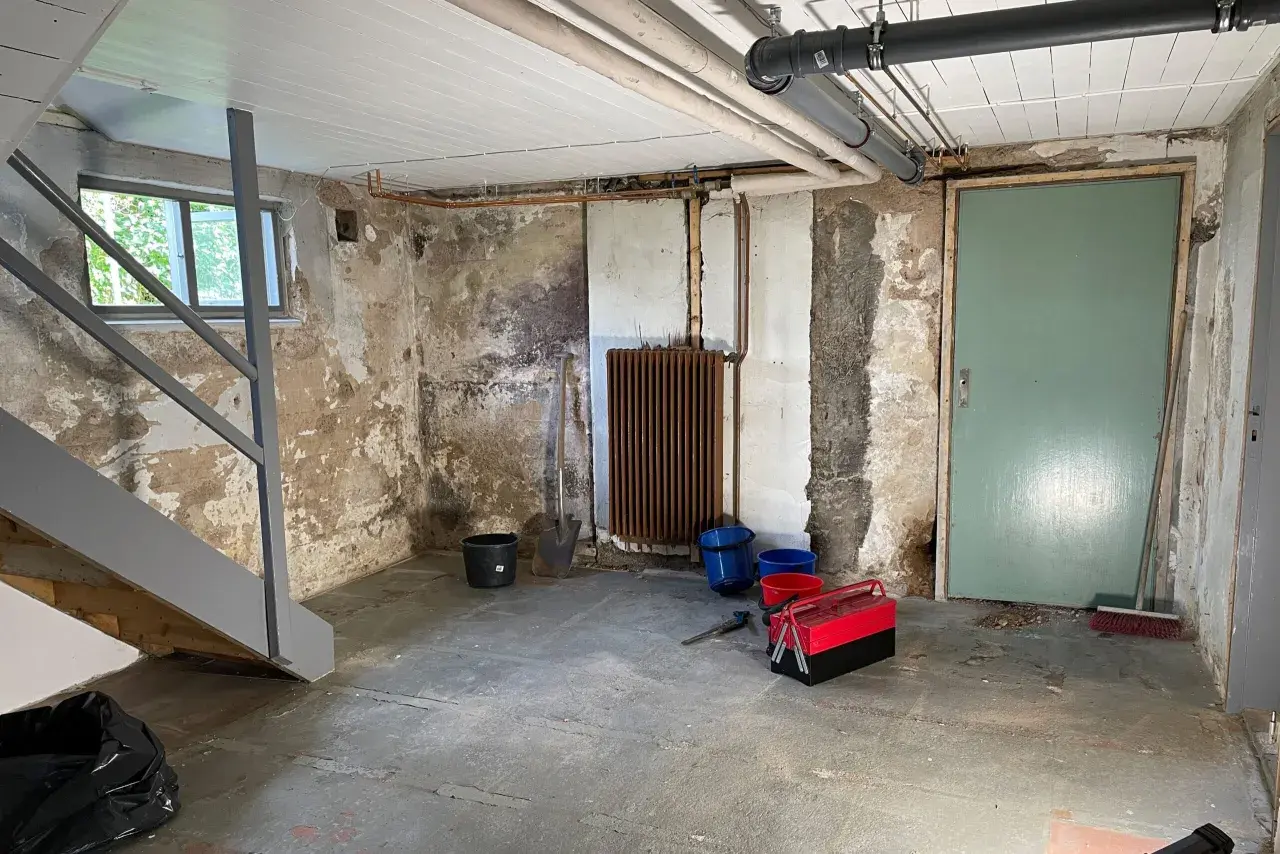 Källare med trappa, fönster, dörr, element och rör. Verktyg och hinkar står på golvet. Förberedelser för FTX ventilation och radonmätning.