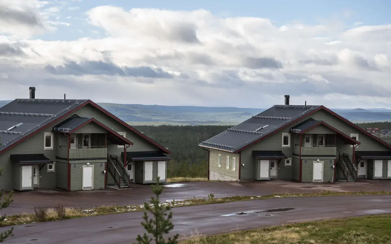 Två moderna knuttimrade stugor i grönt med mörka tak, belägna i ett kuperat landskap med skog och berg i bakgrunden.