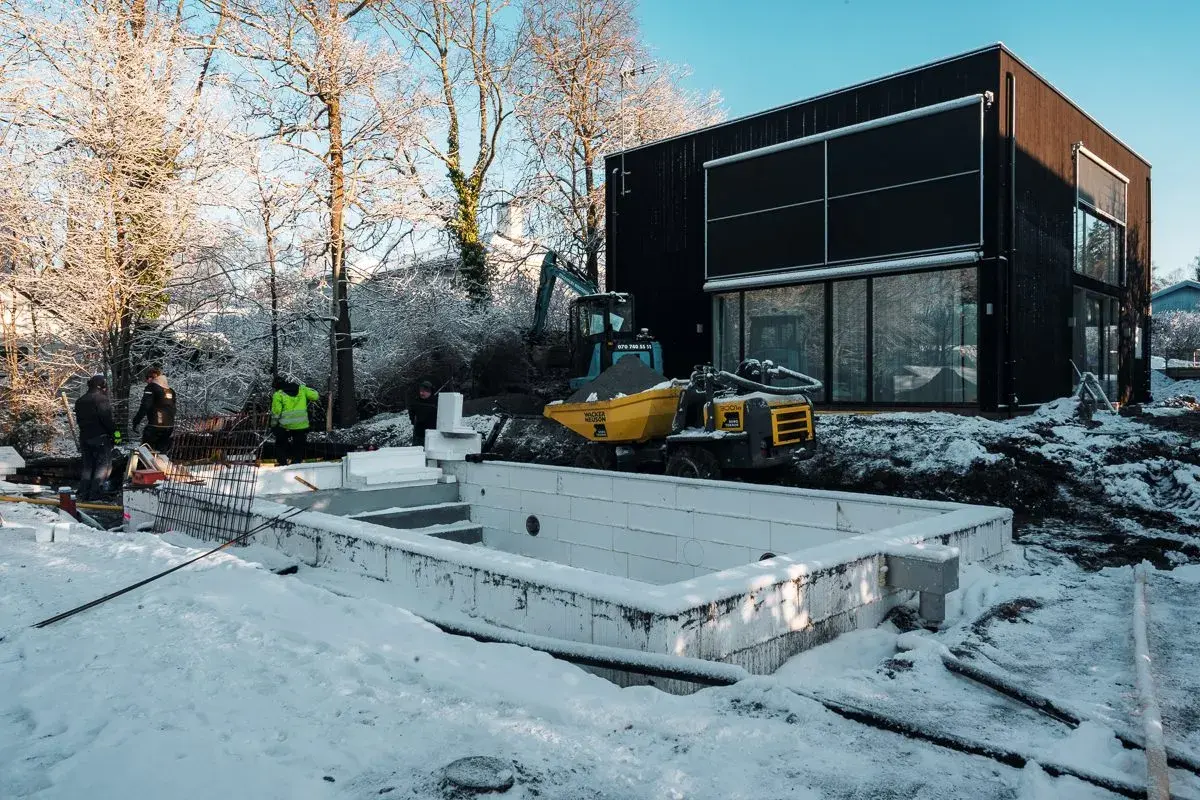 Snö täcker marken vid en pågående utbyggnad av ett hus. En grävmaskin och en tippkärra syns vid en nybyggd grund.