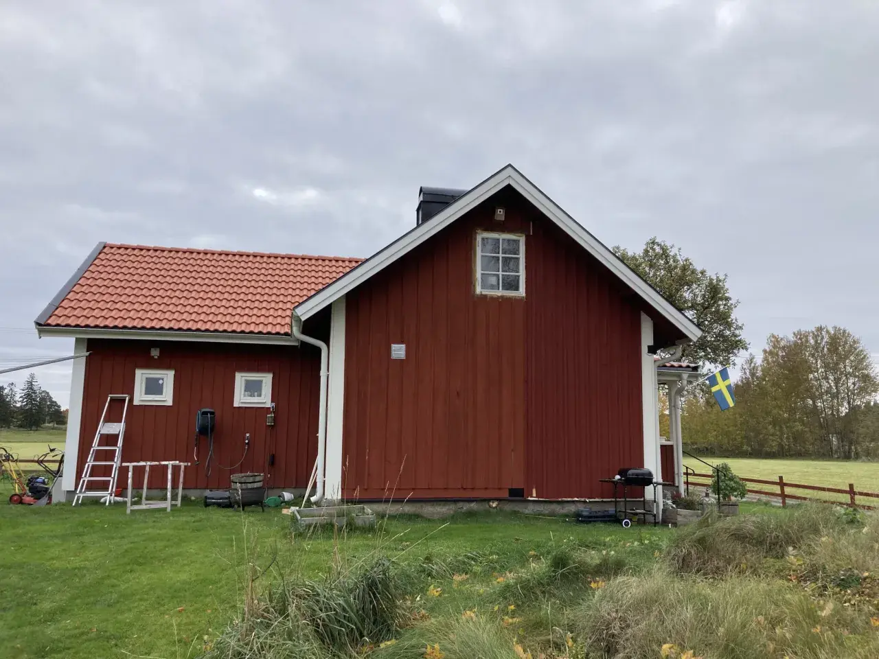 Rött torp med ny tillbyggnad och svensk flagga. Gräsklippare och grill står redo.