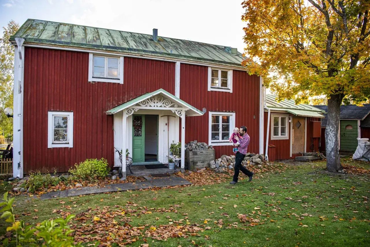 Man bär barn framför ett rött hus från 1700-talet med grönt tak. Höstlöv täcker gräsmattan.
