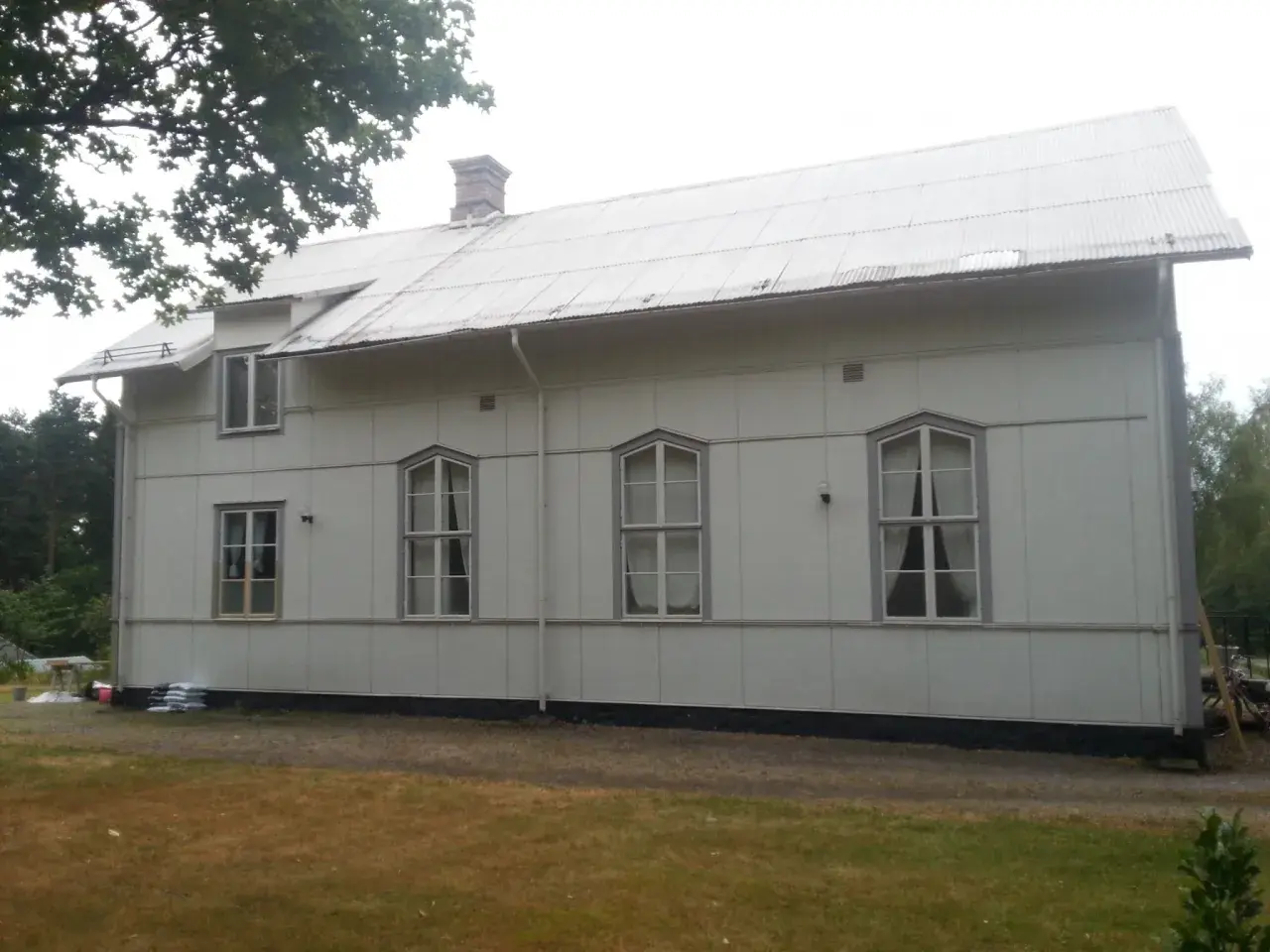 A white building with a corrugated metal roof and several windows, set against a backdrop of trees and a cloudy sky.
