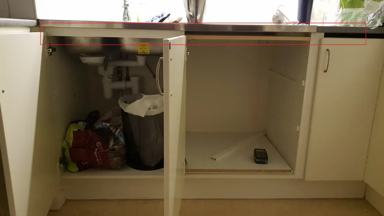 Under diskbänken pågår förberedelser för att installera diskmaskin i skåp. Syns rör, en papperskorg och diverse påsar.