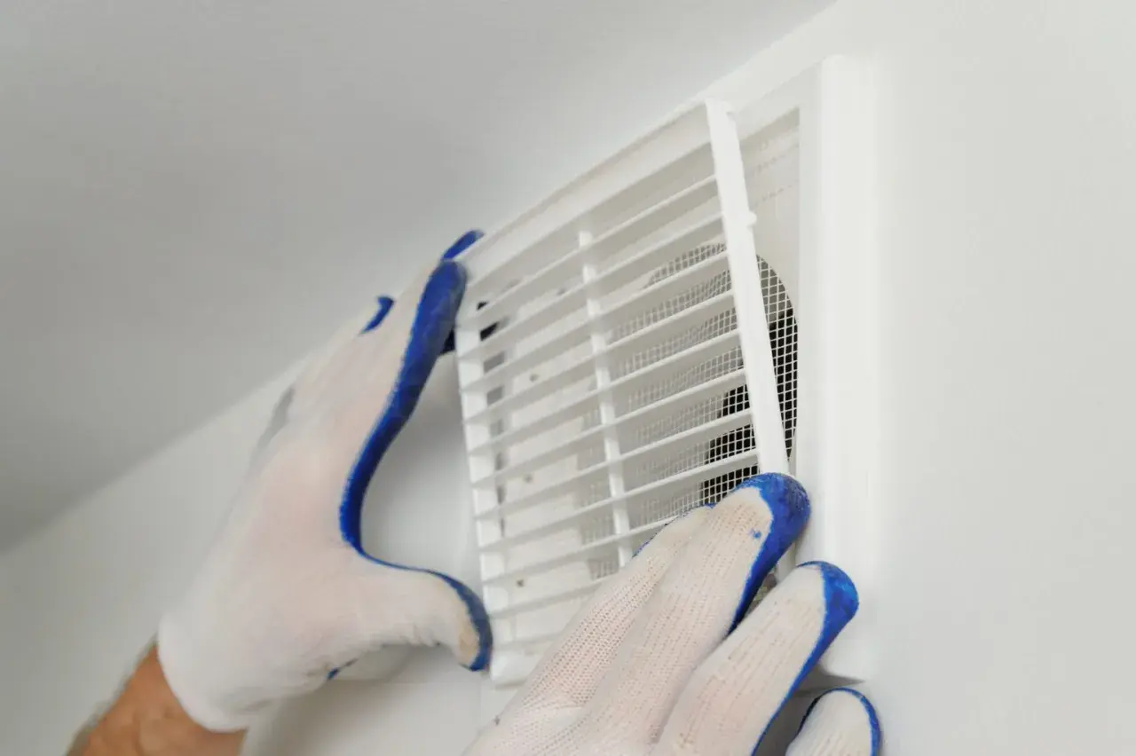 Händer med handskar rengör ett ventilationsgaller för att förbättra dammigt hemma ventilation.