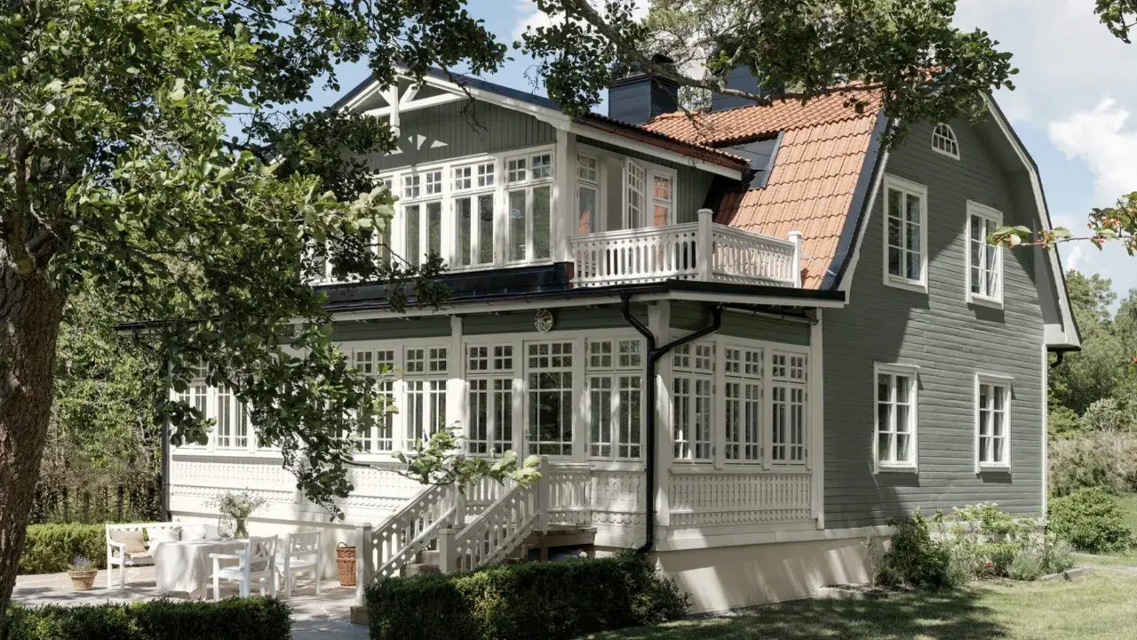 Ett charmigt grått hus med svarta knutar, omgivet av grönska. Ett vackert hus med många fönster och en veranda.