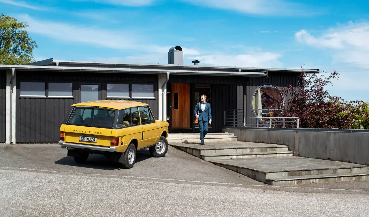 Man i kostym går mot ett funkishus från 60-talet med en gul Range Rover parkerad utanför.