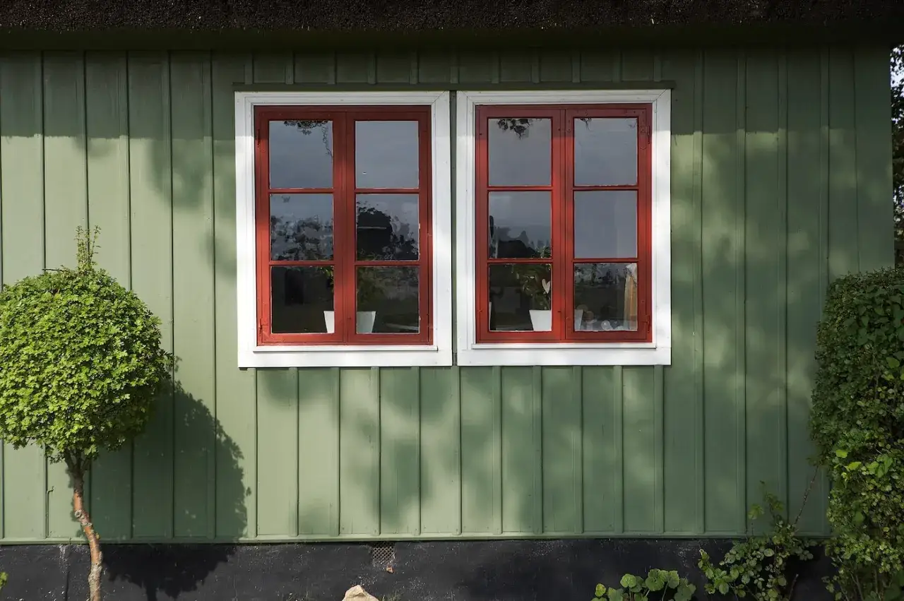 Grönt hus med röda fönsterkarmar. En fin buske står till vänster, redo att måla huset.