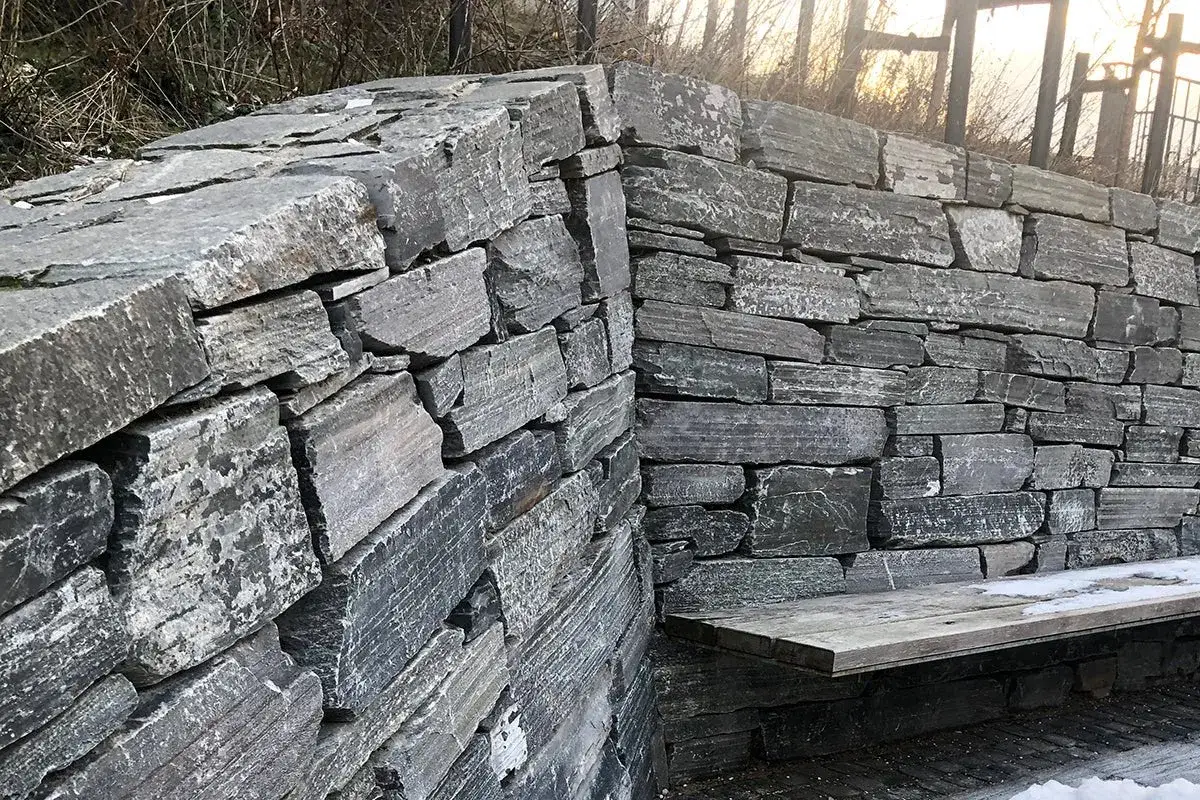 En mur av natursten som ramar in en b&auml;nk. Stenarna &auml;r gr&aring; och oregelbundna, vilket ger ett rustikt intryck.