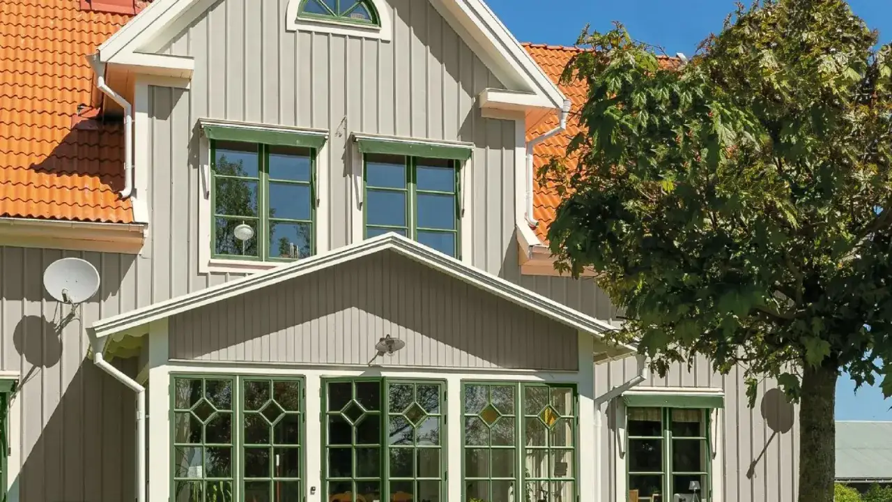 Grått trähus med orange takpannor och gröna fönster. En stor lönn skymtar till höger.