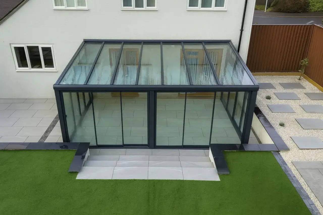 Ett modernt hus med en imponerande glasveranda som sträcker sig ut mot en välskött trädgård med konstgräs och stenplattor.