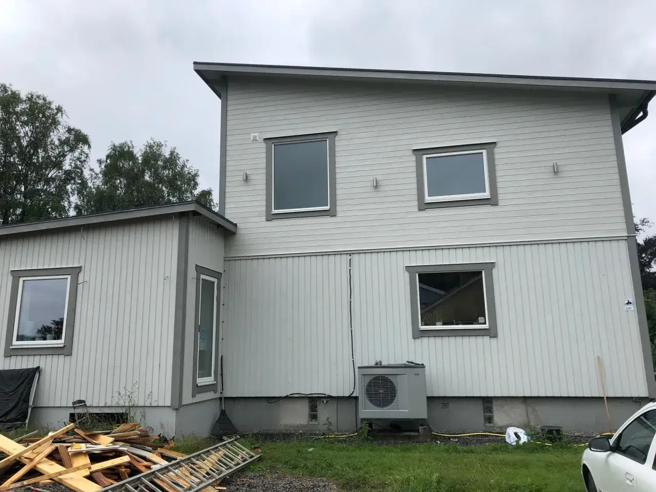 Gr&aring;tt hus med vita f&ouml;nster och en luftv&auml;rmepump. Byggmaterial och en stege ligger p&aring; marken, kanske f&ouml;rberedelser f&ouml;r taklyft pris.