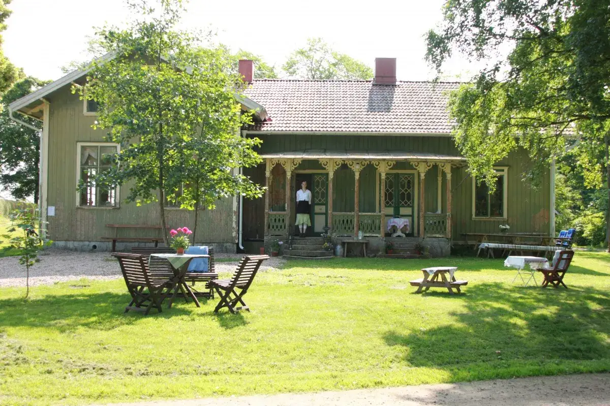 Ett charmigt 1800-tals hus med veranda och trädgårdsmöbler. En kvinna i gammaldags kläder står i dörröppningen.