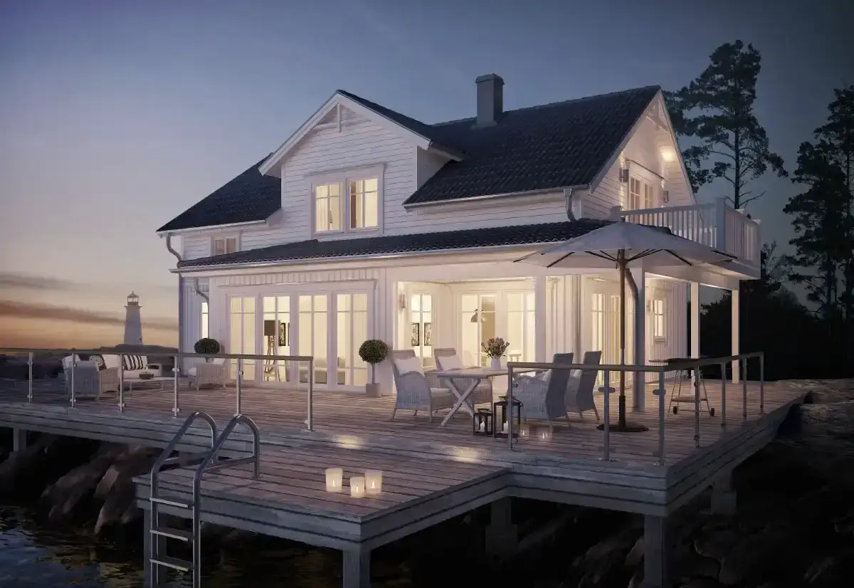 Vitt hus med glasveranda entre vid vattnet, med möblerad altan och en fyr i bakgrunden.