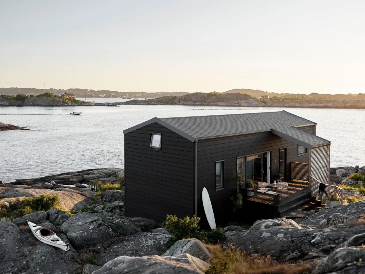 Ett modernt, m&ouml;rkt gr&aring;tt hus med svarta knutar st&aring;r p&aring; en klippig kust. En kajak ligger p&aring; stenarna vid vattnet.