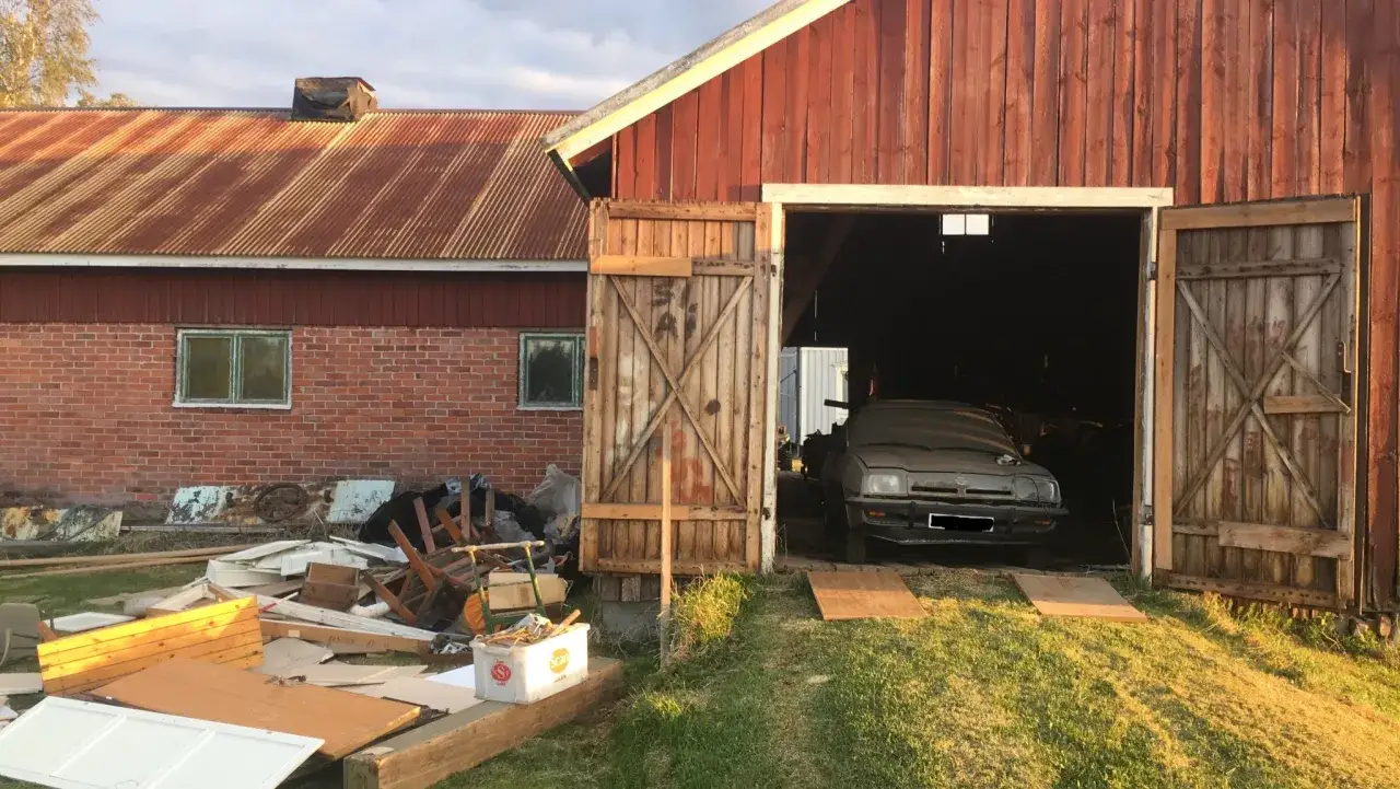 Vackra ladugårdar med en gammal bil i garaget. Runtomkring ligger bråte och byggmaterial.