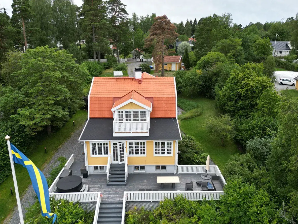Gult hus med orange tak och en takkupa, omgivet av gr&ouml;nska. En altan med m&ouml;bler och en svensk flagga. En utbyggnad av taket har gjorts.