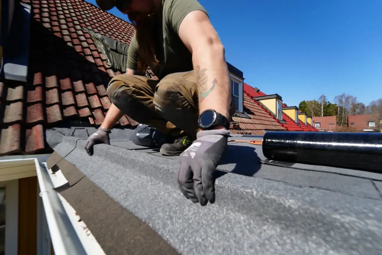 Man med sk&auml;gg och tatueringar arbetar p&aring; ett tak, l&auml;gger papp. Solen skiner och himlen &auml;r bl&aring;.