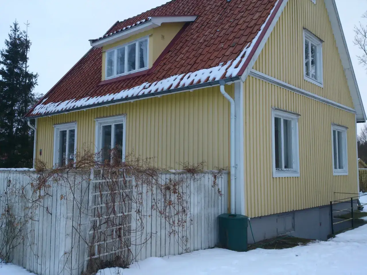 Gult 40-talshus med tegeltak och snö på takfoten. Ett staket med klätterväxter löper framför huset. Planer på att renovera 40-talshus?