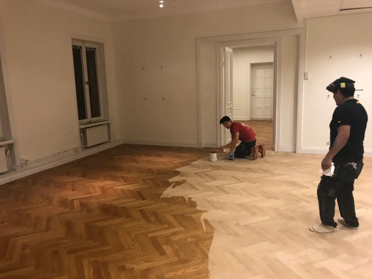 Två män arbetar med att slipa och lacka ett parkettgolv. Halva golvet är mörkt och lackat, medan den andra halvan är ljus och nybehandlad.