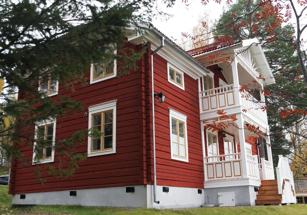 Röd knuttimrad stuga med vita detaljer och balkonger. Träd med höstfärger ramar in huset.