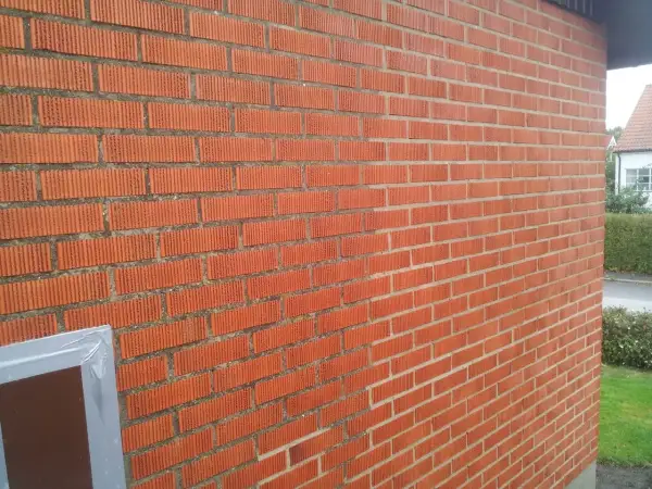En röd tegelfasad med vertikala spår i tegelstenarna. En del av en fönsterkarm syns i nedre vänstra hörnet.