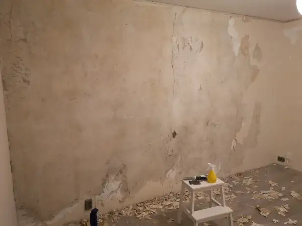 Förberedelser för att tapetsera på betong. Gamla tapeter skrapas bort från en sliten vägg. Verktyg och en sprayflaska står på en pall.
