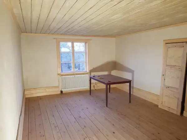 Ett rum med kalkputs inomhus, trägolv och en takbjälke. Ett fönster med utsikt över träd och ett bord med stolar.