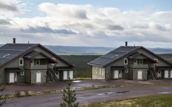 Två moderna knuttimrade stugor i grönt med mörka tak, belägna i ett kuperat landskap med skog och berg i bakgrunden.