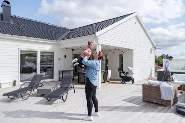 Kvinna lyfter barn på altanen framför ett av de färdiga husen. Barnvagn och utemöbler syns också.