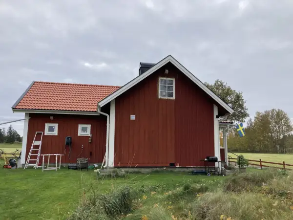 Rött torp med ny tillbyggnad och svensk flagga. Gräsklippare och grill står redo.