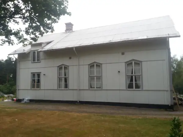 A white building with a corrugated metal roof and several windows, set against a backdrop of trees and a cloudy sky.