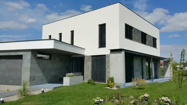 Modernt hus med vit, grå putsad fasad och stenbeklädda partier. Grönskande trädgård och garage.