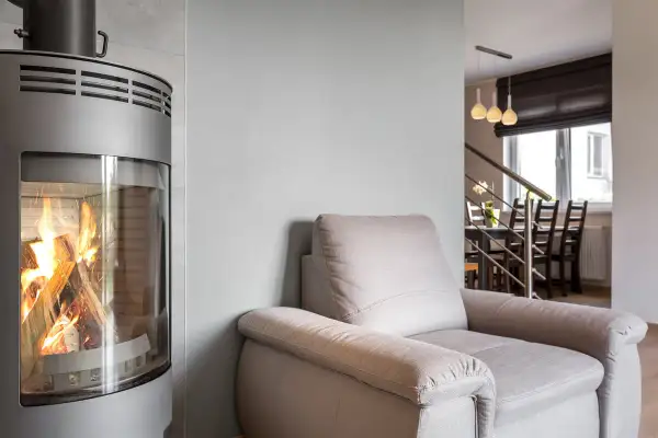 En modern kamin med brinnande ved. En bekväm fåtölj står bredvid, perfekt för en mysig kväll. Kanske dags att renovera kakelugn för en liknande känsla?