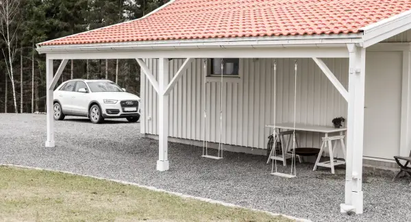 Vit bil parkerad vid en vit träfasad med röd tegeltak. En utbyggnad hus långsida med hängande gungor och ett bord.