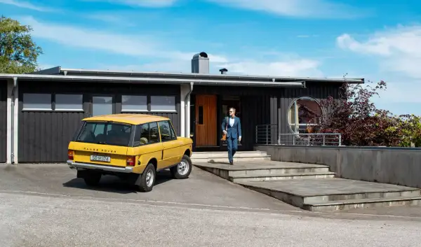 Man i kostym går mot ett funkishus från 60-talet med en gul Range Rover parkerad utanför.